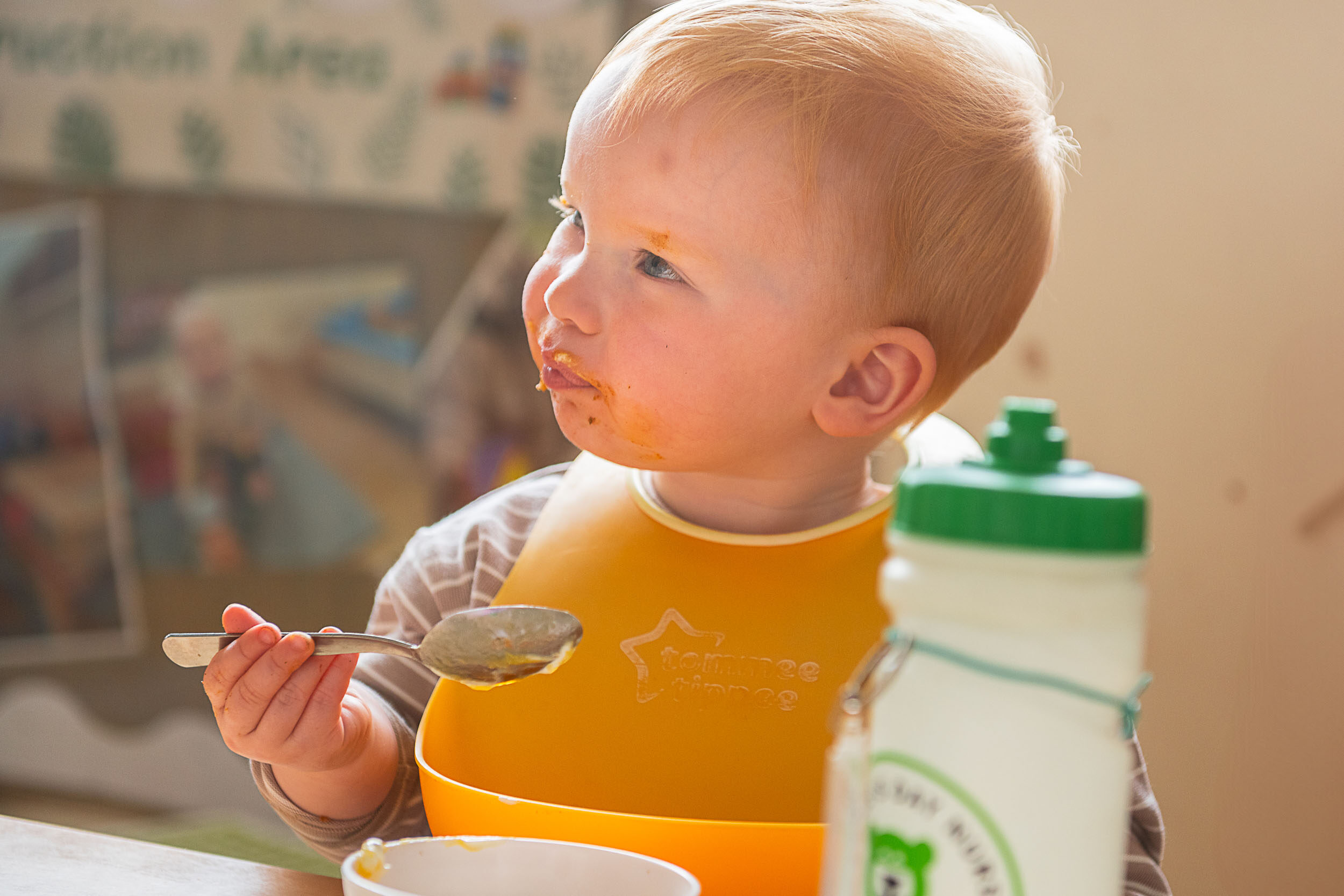 young child wearing a bib, eating with a spoon, face covered in sauce