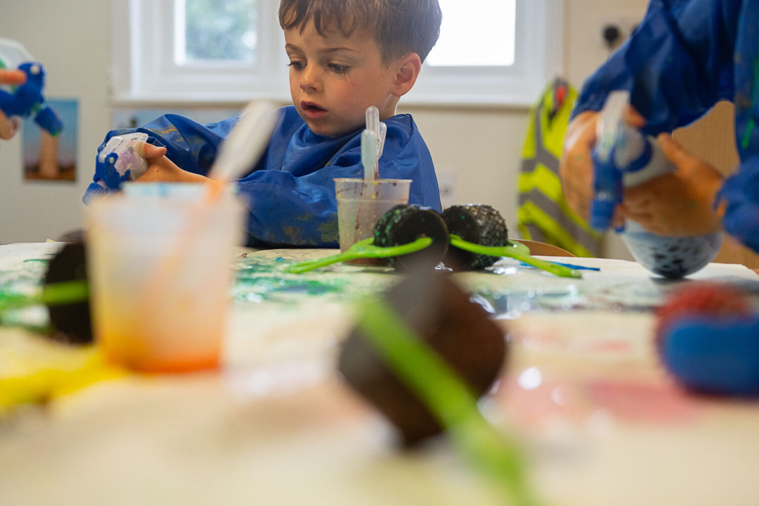 Table of paints and paint rollers, little boy spraying a bottle of paint onto the table