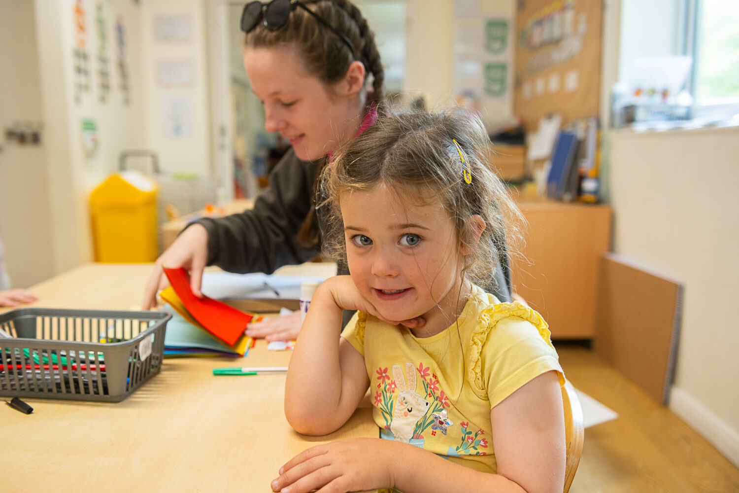 Little girl sat with a member of staff doing arts and crafts. the little girl is posing at the camera with her hand under her chin
