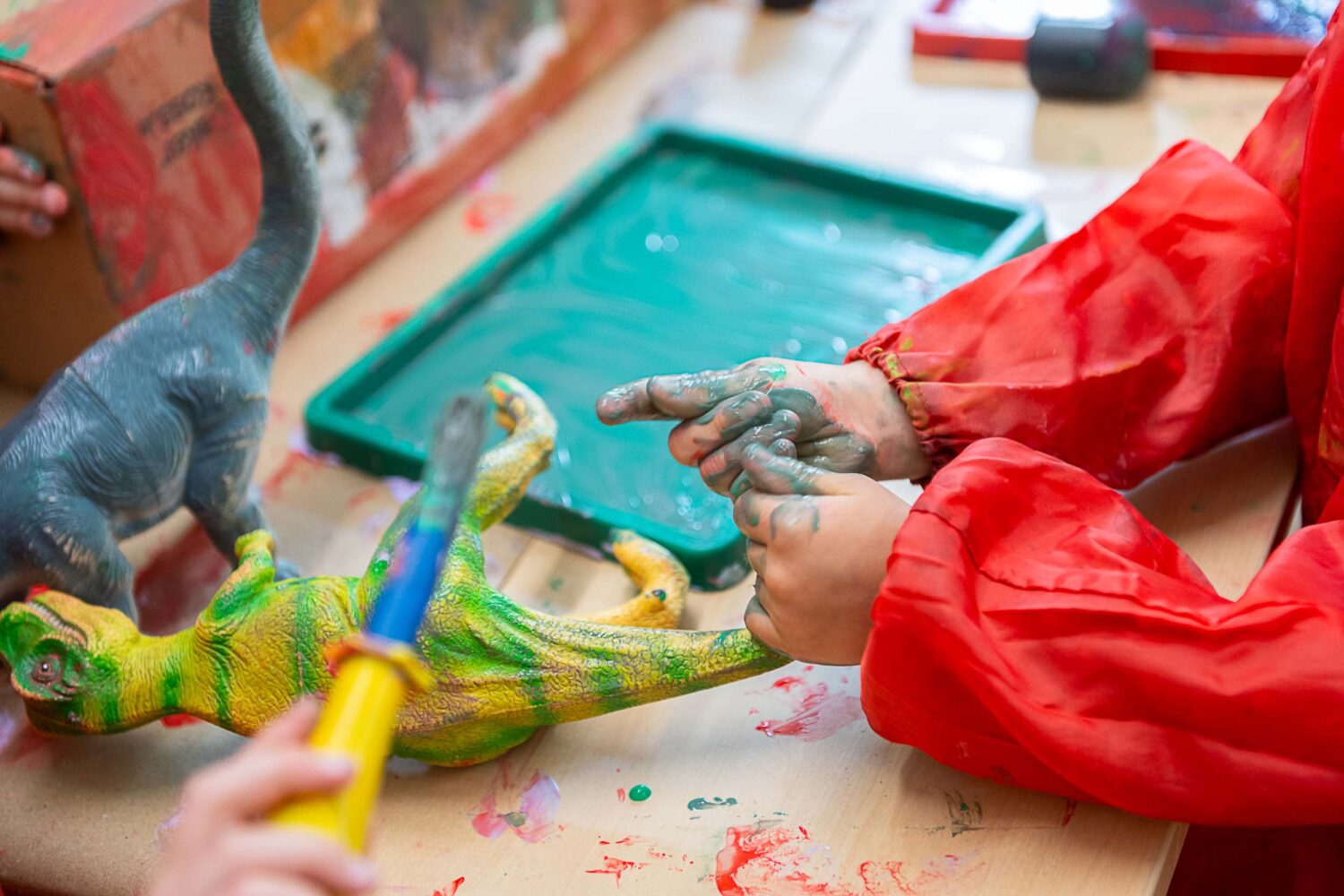 A young child with their hands covered in paint, rubbing paint onto a toy dinosaur