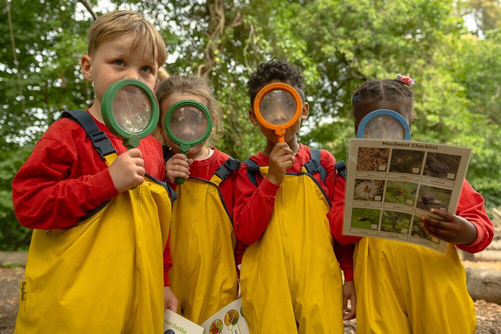 Group of children holding magnifying glasses over their faces and wearing waterproofs, one is holding a information sheet