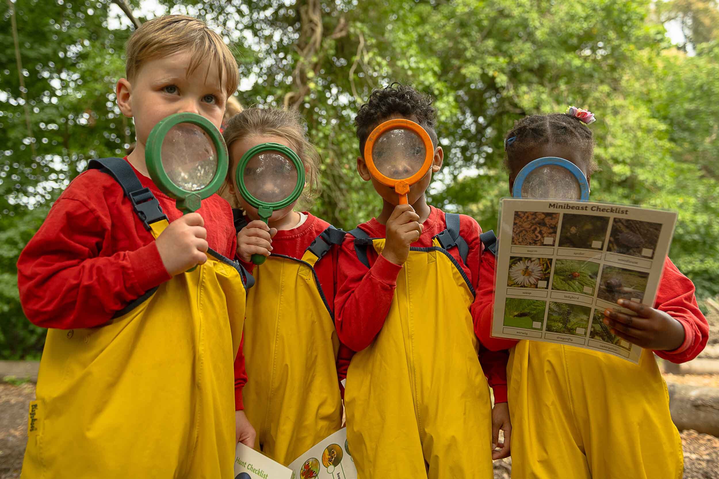 Group of children holding magnifying glasses over their faces and wearing waterproofs, one is holding a information sheet