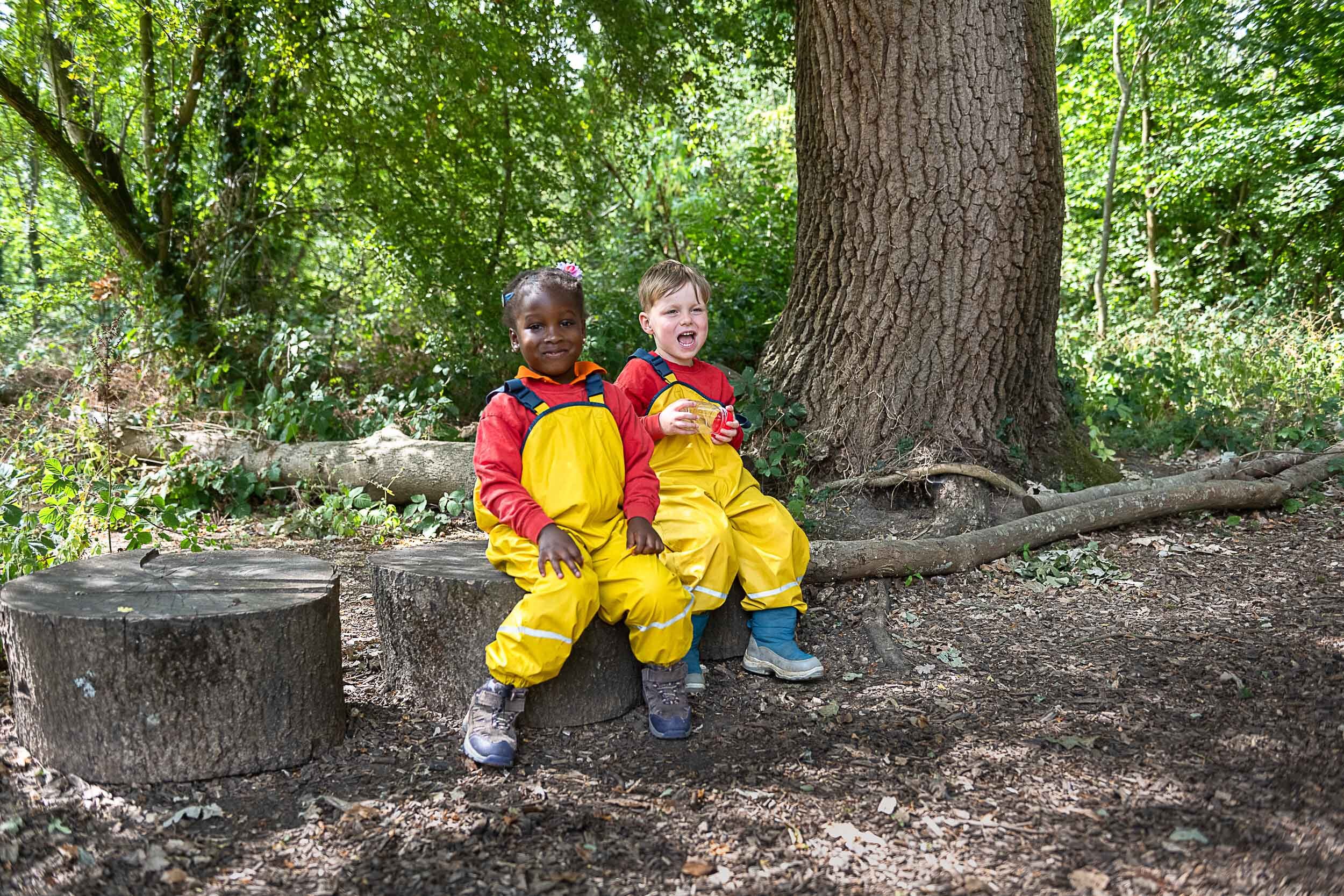 two children in their outdoor waterproofs, sat on tree stumps, smiling