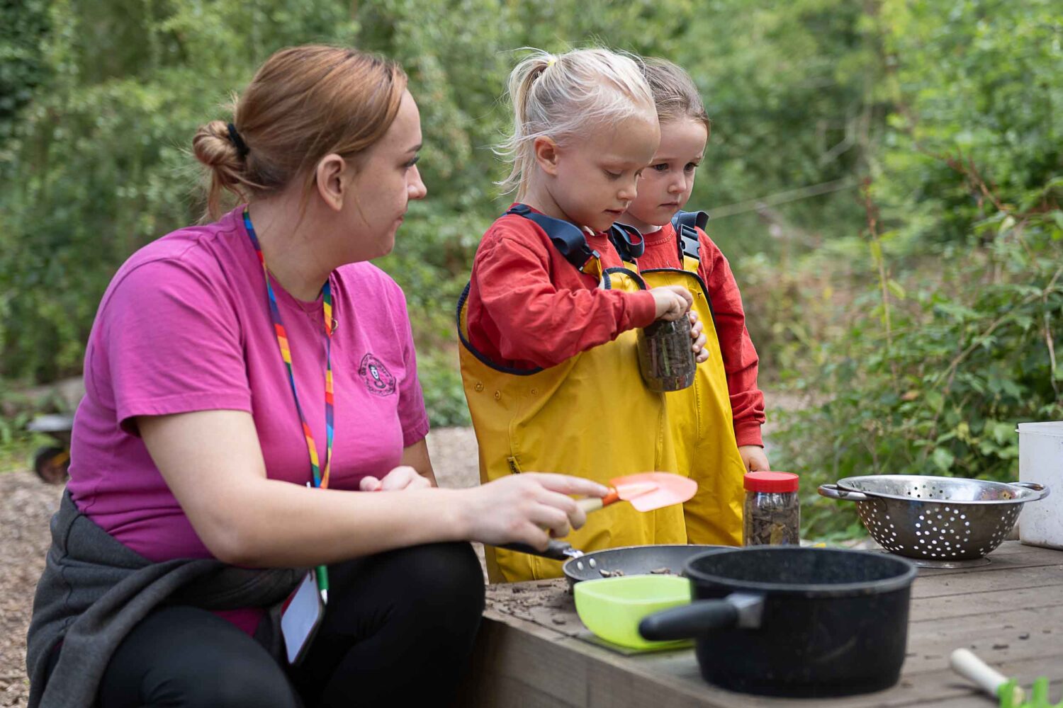 A member of staff playing pots and pans with two little girls in the forest school