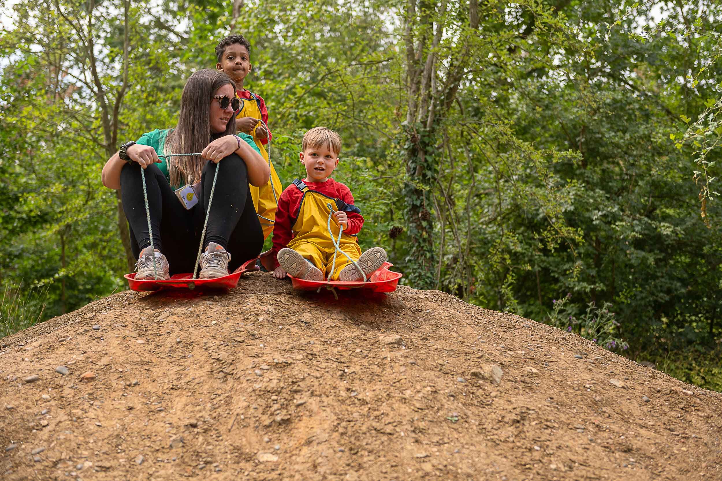 A member of staff with two children on top of a dirt mound. Both are sitting on sledges waiting to slide down the mound